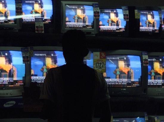 man stands before rows of television screens all showing the same image of the burning Twin Towers of the WTC