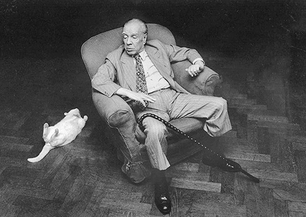 photo of Jorge Luis Borges seated in a chair with white cat stretching on the floor nearby