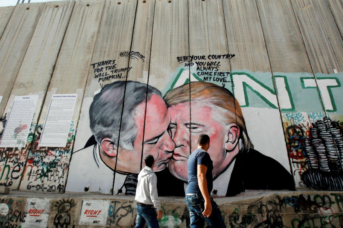 Palestinian men walk past a graffiti by street artist Lushsux, depicting US President Donald Trump kissing Israeli Prime Minister Benjamin Netanyahu drawn on the controversial Israeli separation barrier separating the West Bank town of Bethlehem from Jerusalem, on October 29, 2017. (Photo credit: MUSA AL SHAER/AFP via Getty Images)
