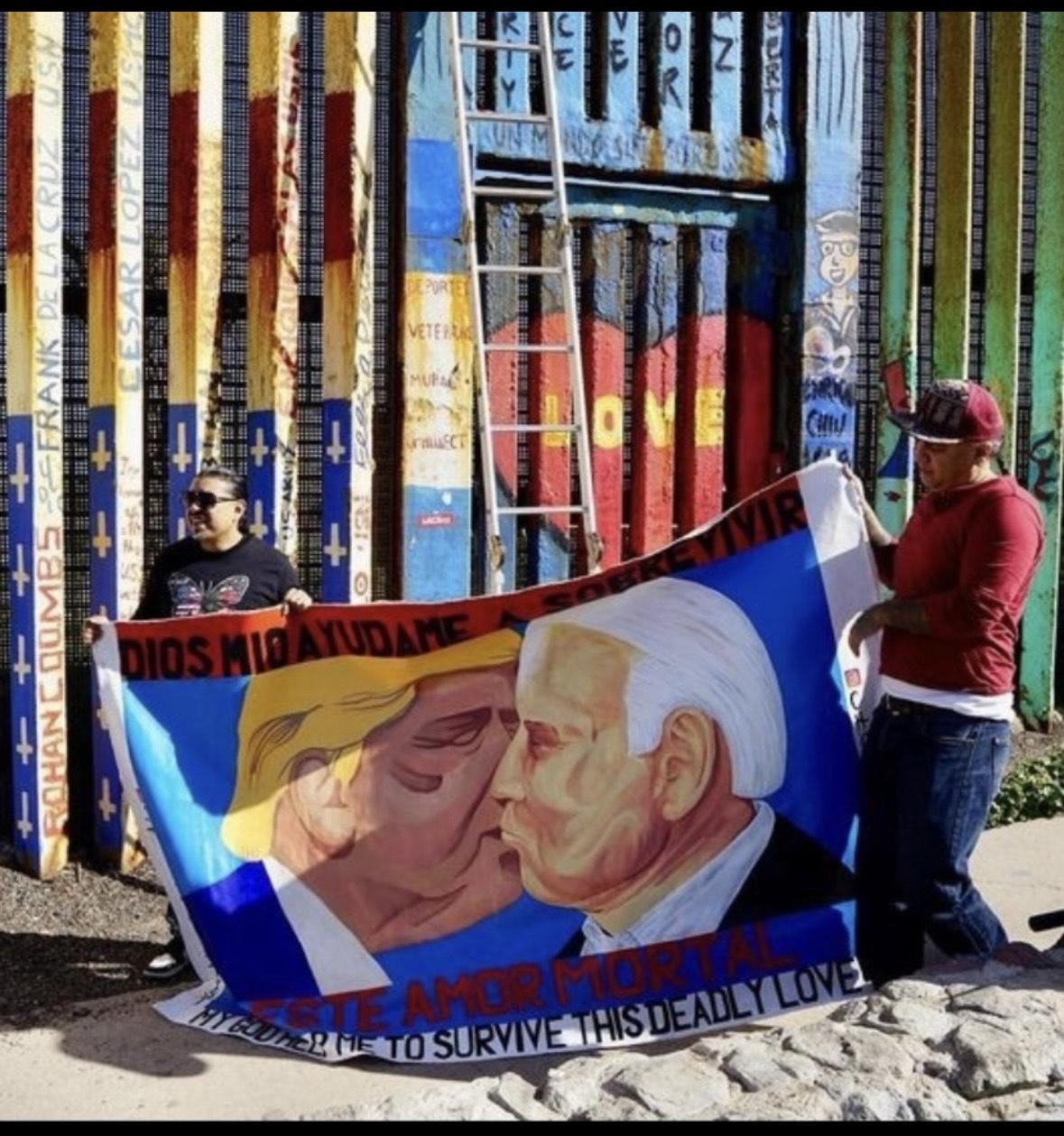 Artists Javier Salazar Rojas (left) and Chris Cuauhtli (right) hold their mural portraying a symbolic kiss between U.S. Presidents Donald Trump and Joe Biden at the border wall in Friendship Park, Tijuana, Mexico in March 2023. (Photo credit: JAVIER SALAZAR ROJAS / @DeportedArtist)