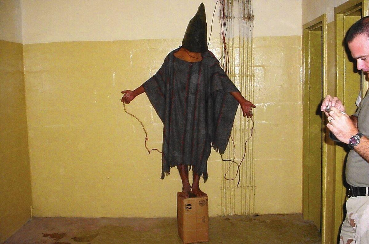 shrouded prisoner at Abu Ghraib prison in Iraq stands on box, electrical wires attached to hands wires
