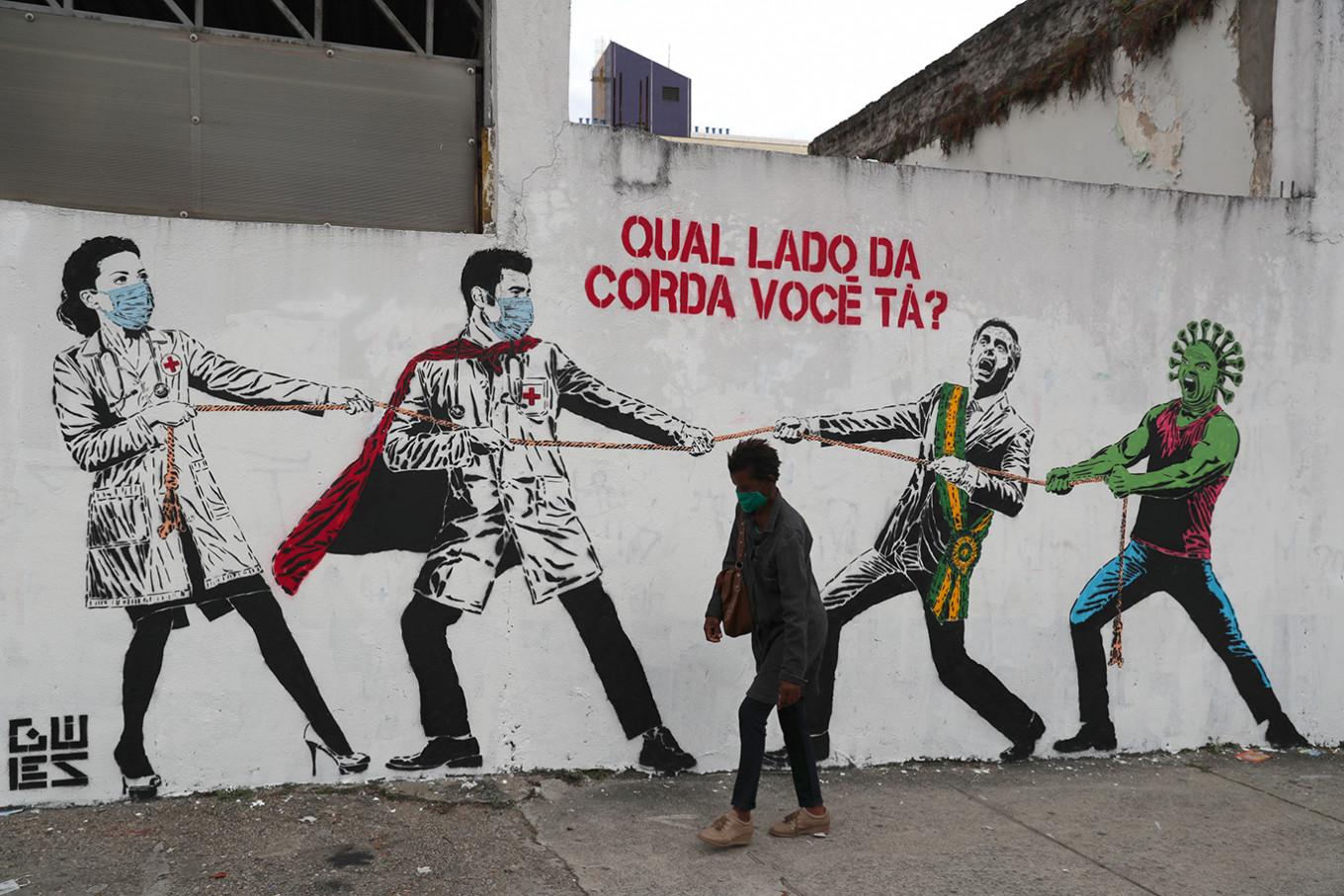 a woman walks by a mural depicting Jair Bolsonaro and a personified COVID-19  in a tug of war against doctors