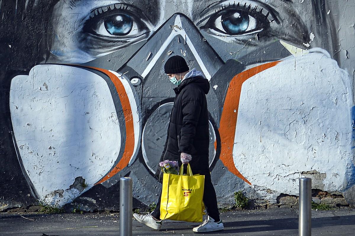 A man wearing a face mask walks with his groceries past a mural depicting a woman wearing a gas mask in Milan, Italy.