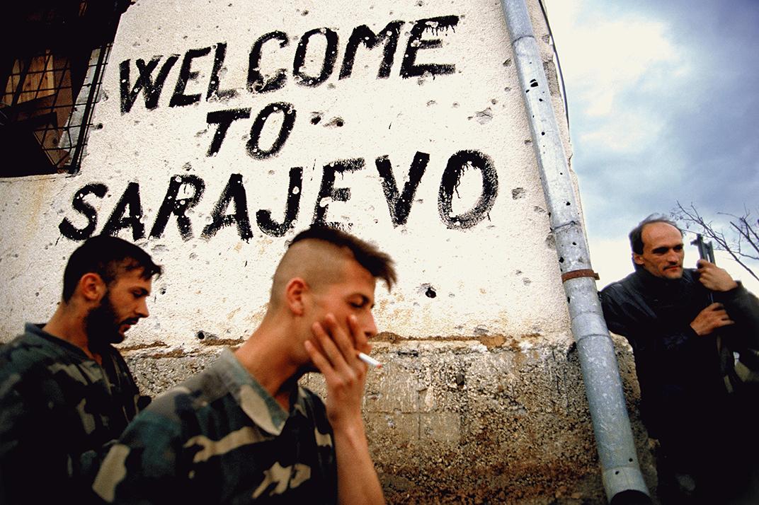 soldiers walk past bullet-riddled street corner on which is painted 'Welcome to Sarajevo'