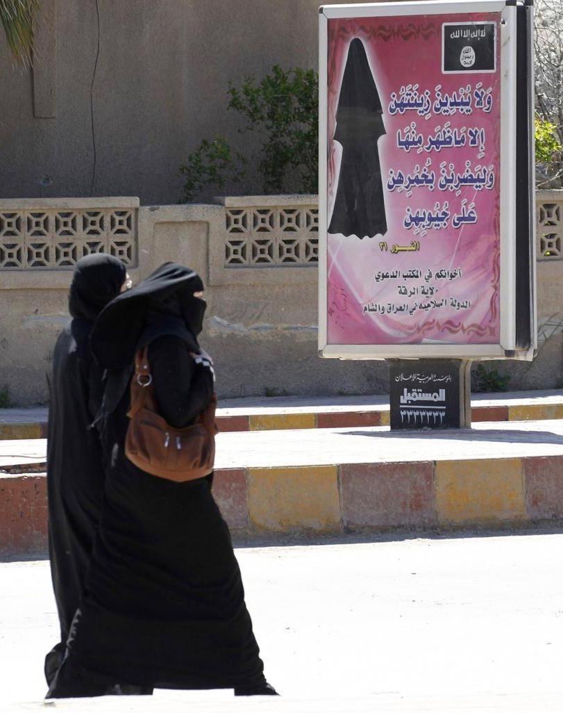 veiled women walk past billboard with verse from the Qur’an urging women to wear a hijab. Raqqa, Syria, 2014. Reuters