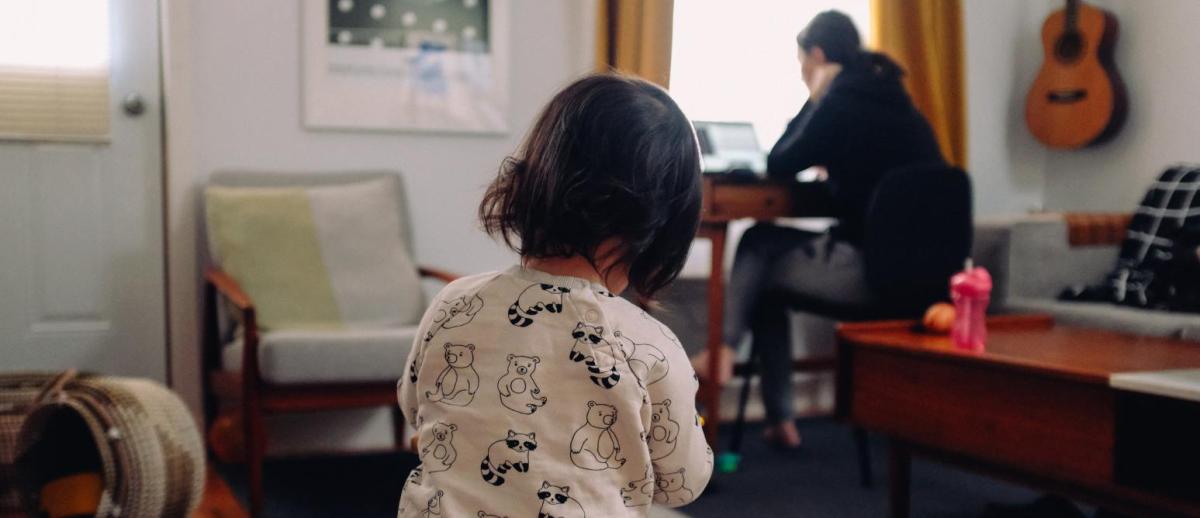 A woman with laptop works at a small desk while a child plays with toys in the foreground