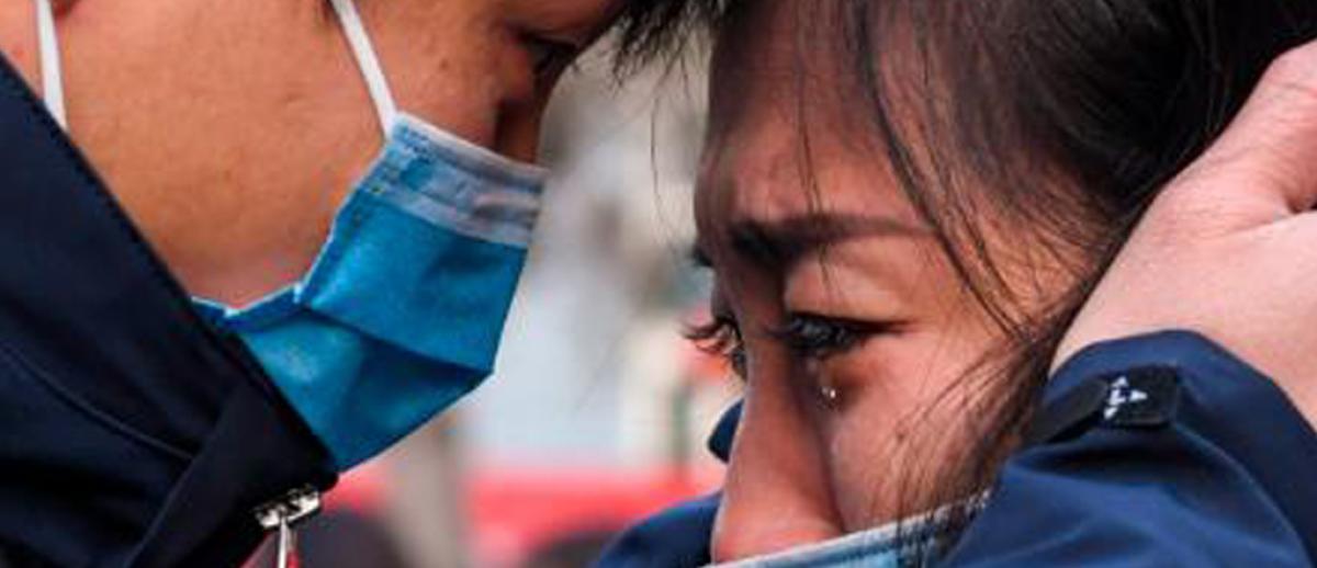 a man consoles a weeping woman, both wearing surgical masks