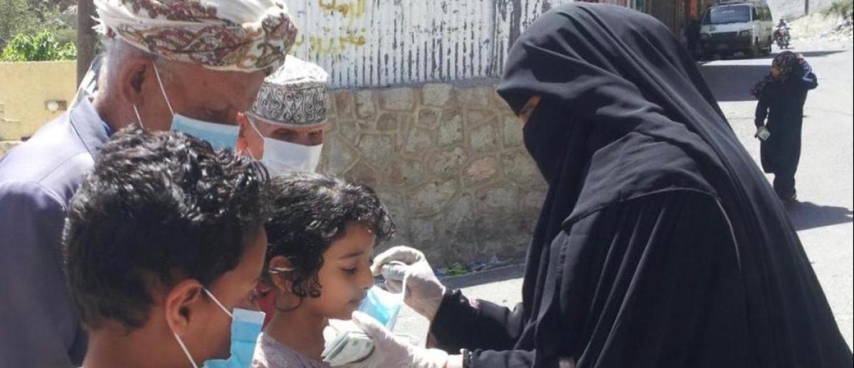 a woman in black burqa and gloves helps children put on surgical facemarks in unidentified Yemeni town