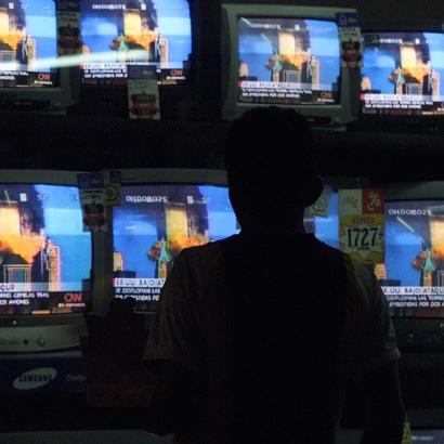 man stands before rows of television screens all showing the same image of the burning Twin Towers of the WTC